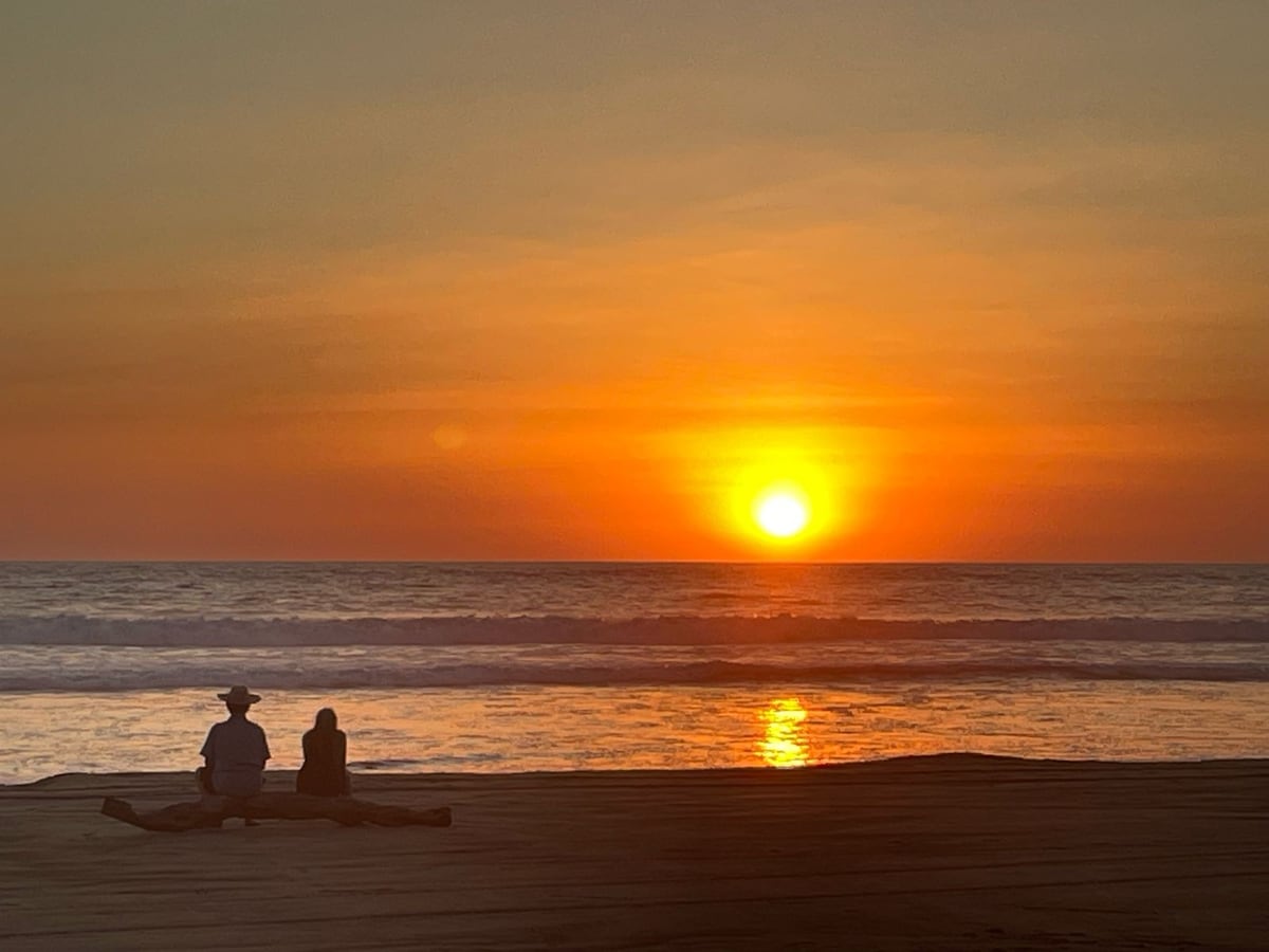 A serene beach scene captures two individuals seated on the sand, gazing at a vibrant sunset. The sky transitions through warm hues of orange and yellow as the sun sets over the calm ocean, reflecting softly on the water's surface.