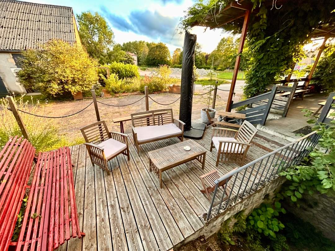An outdoor seating area is presented, featuring wooden furniture arranged on a wooden deck. The space is surrounded by greenery and flowers, with a vibrant red bench on one side. A pathway and neighboring greenery are visible in the background.