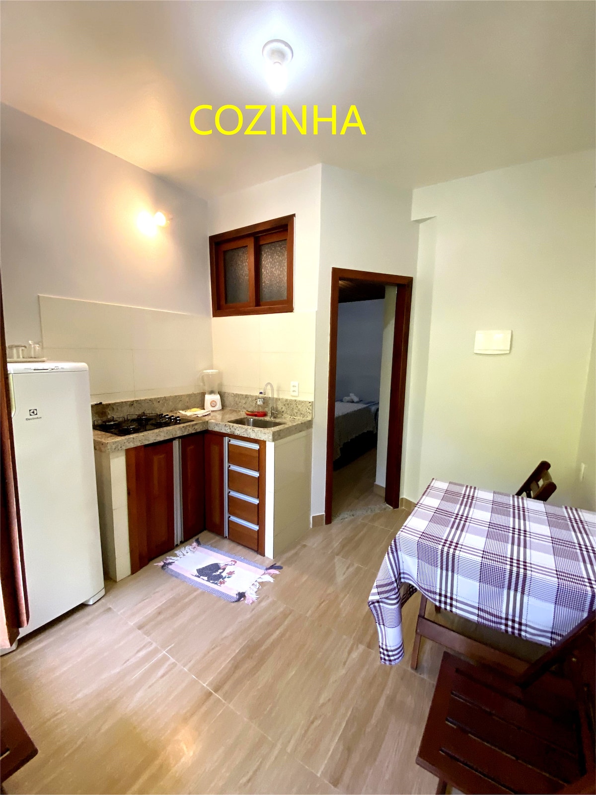 The kitchenette features wooden cabinetry and a granite countertop, containing a cooktop, refrigerator, and various kitchen utensils. A small dining table with wooden chairs is positioned nearby, and natural light fills the space through a window, enhancing the functionality of the area.