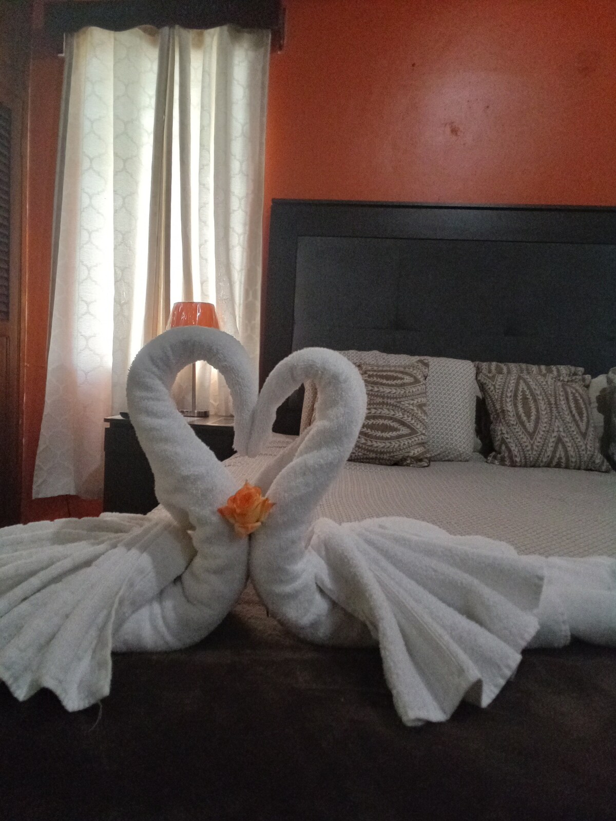 Two towel swans elegantly shape a heart in the foreground, placed on a dark surface. Behind them, a spacious bed is situated against a textured orange wall, framed by soft curtains, and accompanied by patterned pillows.