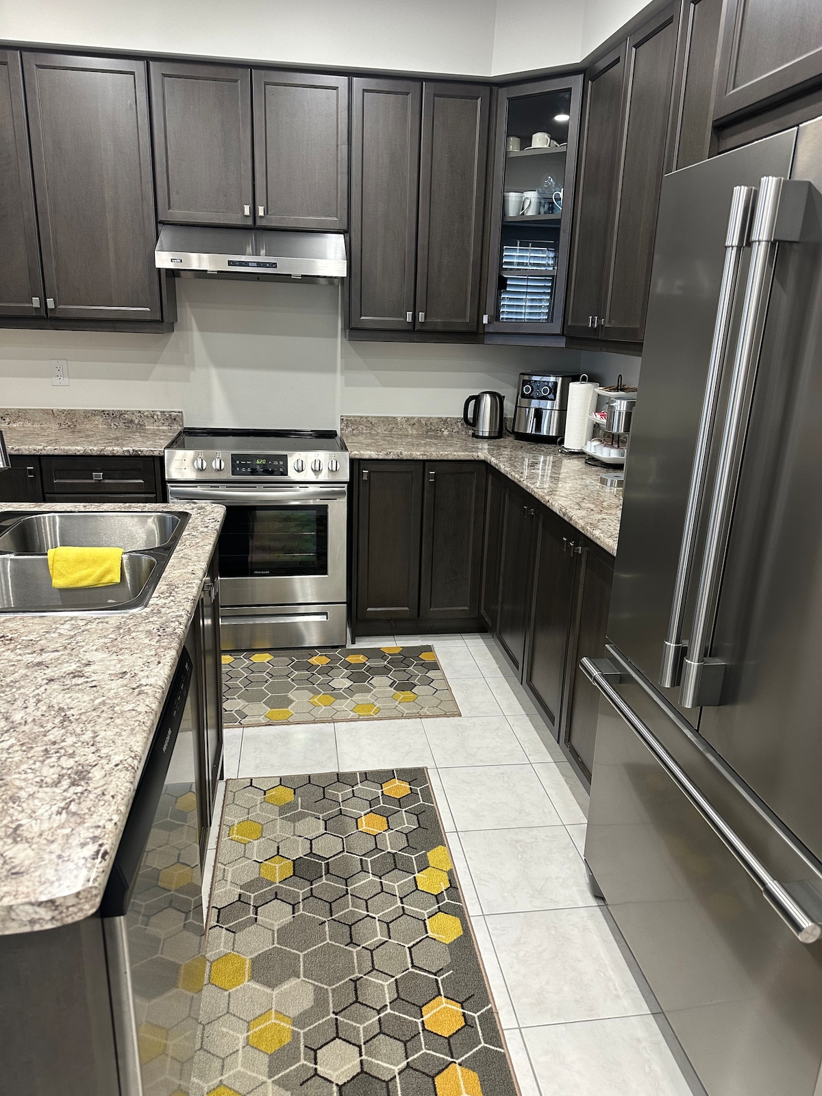 A modern kitchen features dark wood cabinetry, stainless steel appliances, and granite countertops. Brightly patterned rugs add color to the floor. The layout includes a double sink and a stove, with various kitchen appliances organized on the countertop.