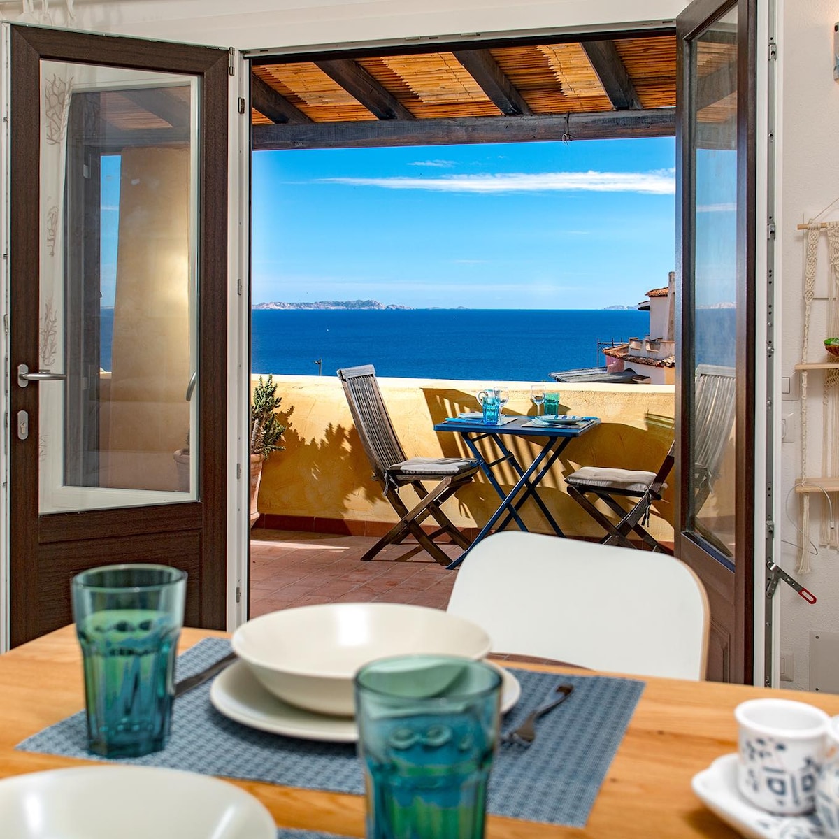 An inviting view opens from the dining area through glass doors, showcasing a terrace with a small table and chairs overlooking the sea. The setting includes wooden beams on the ceiling and a table adorned with plates and glasses, creating a bright, airy atmosphere.