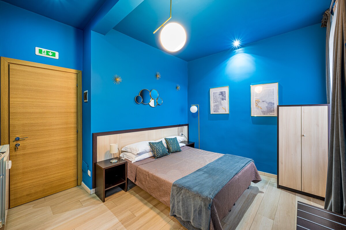 A double room features a spacious bed with a neutral-colored blanket. A vibrant blue wall is accentuated by two framed artworks and wall decor. Soft lighting is provided by a modern ceiling fixture and bedside lamp. A cabinet stands next to the bed.