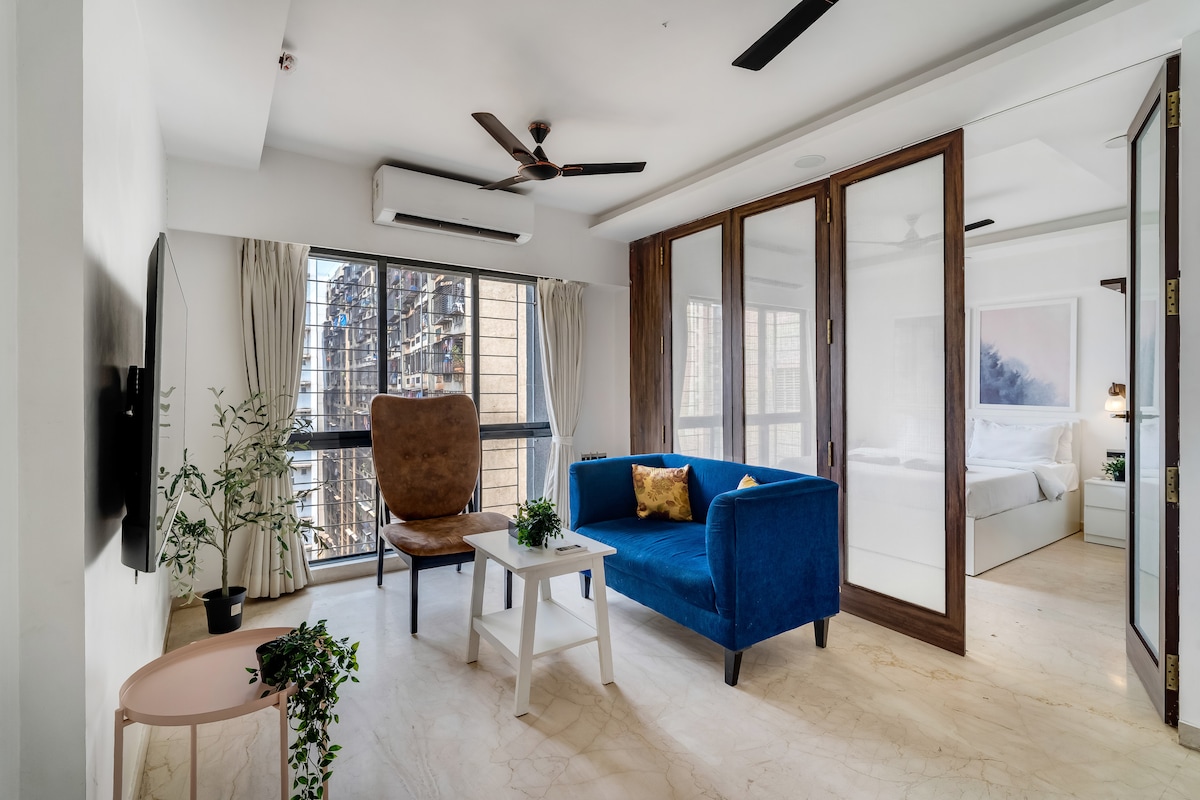 The living area features a blue sofa and a stylish armchair, complemented by a compact white coffee table. Large windows provide ample natural light and views of the surroundings. The space is designed for a modern aesthetic, with plants adding a touch of greenery.