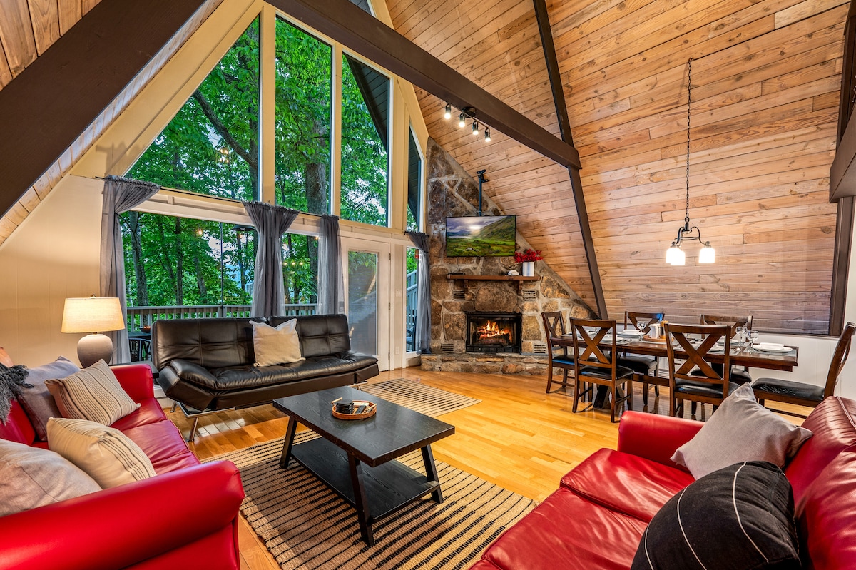 An open living area showcases a high A-frame ceiling with wood accents and large windows offering views of surrounding trees. Cozy seating arrangements include a pair of red sofas, positioned near a stylish coffee table. A stone fireplace and a dining area with dark wood chairs are visible in the background.
