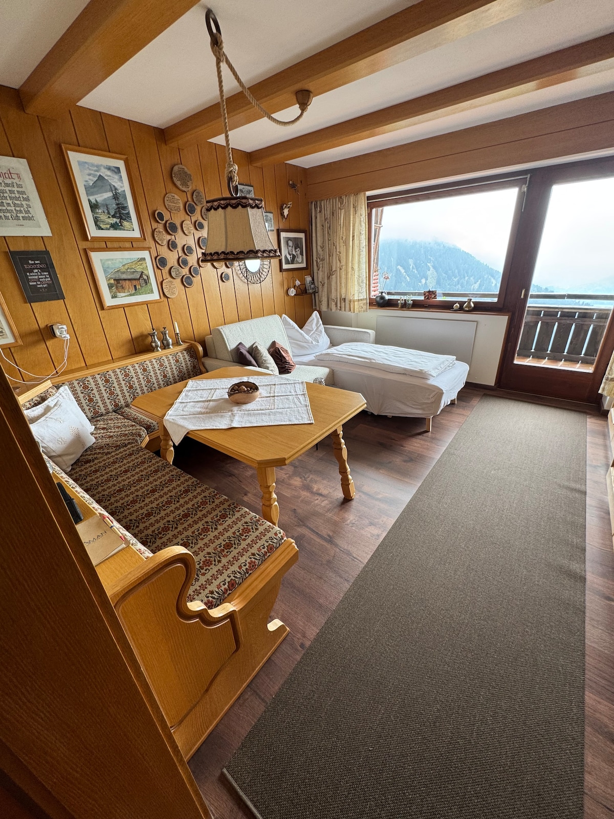 The living area features a wooden table surrounded by a cozy bench and two armchairs, with mountain views presented through a large window. A neutral area rug covers the floor, and several decorative items, including artwork on the walls, contribute to a warm, welcoming environment.