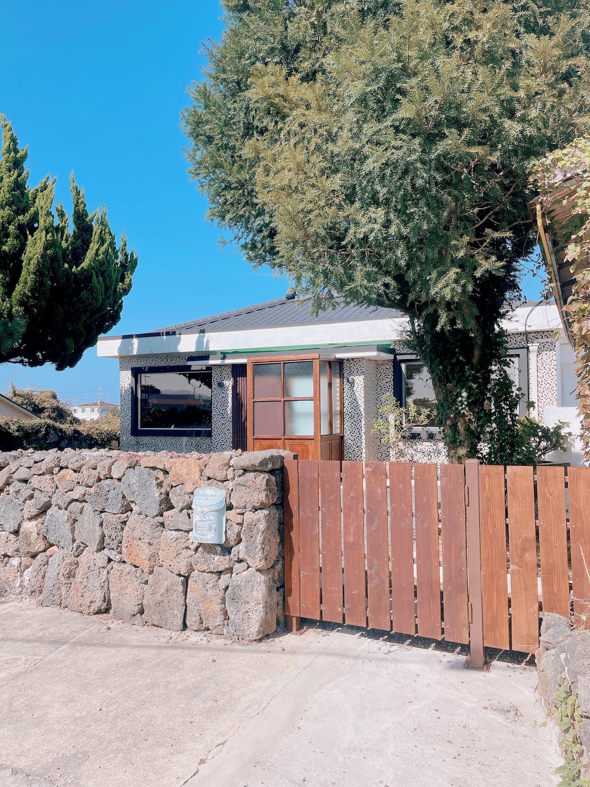 A welcoming exterior features a stone fence and wooden gate, leading to a single-story house surrounded by greenery. Tall trees provide shade, and a well-maintained facade showcases a blend of modern and traditional architectural elements.