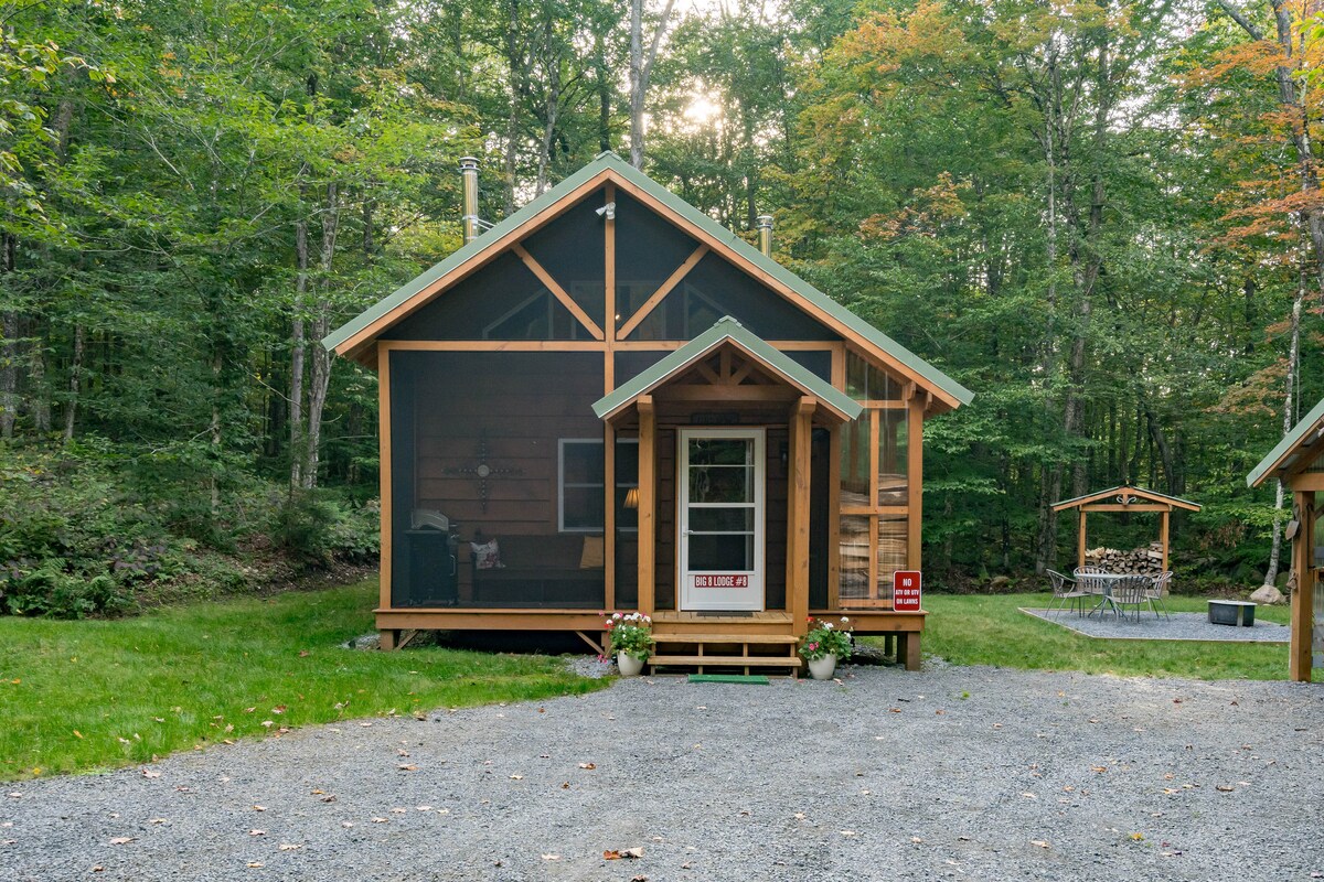 A charming cabin is nestled among trees, showcasing a screened-in front porch with an entrance door and flower pots beside the steps. The exterior features natural wood siding and a gabled roof, blending harmoniously with the surrounding landscape.