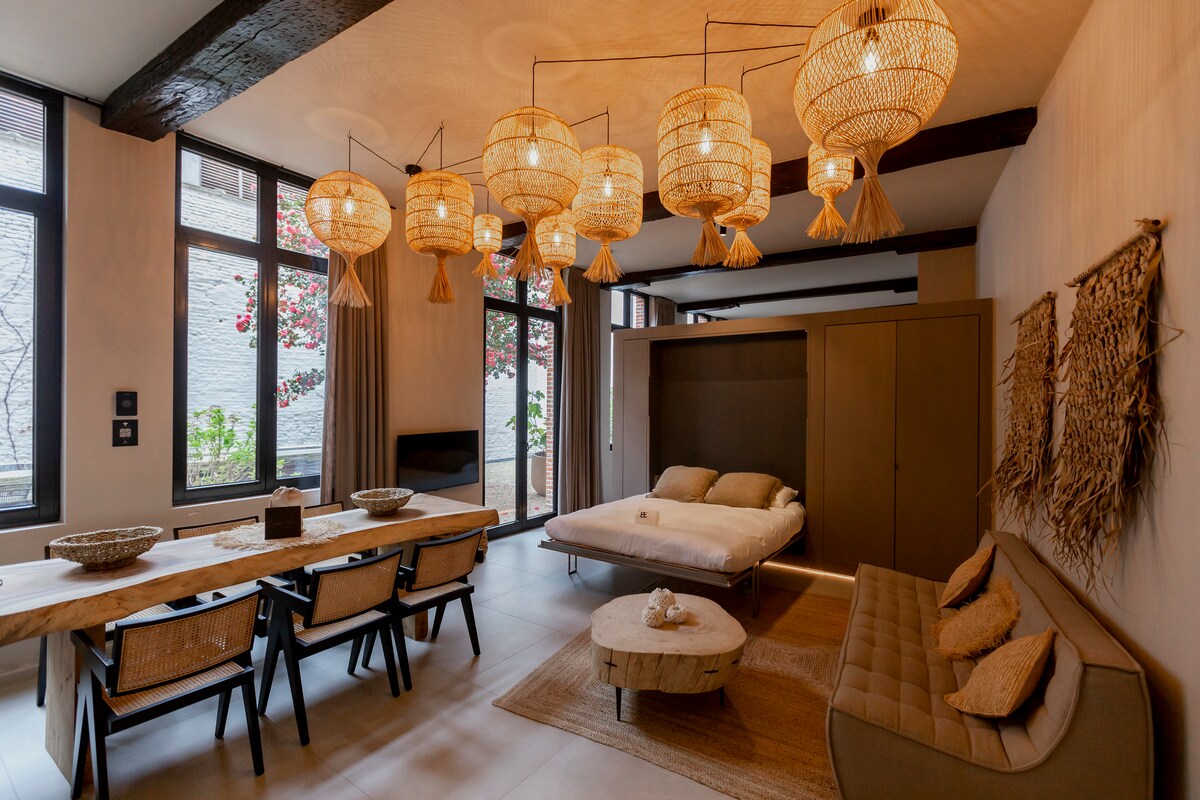 A spacious living area features multiple woven pendant lights hanging from a wood-beamed ceiling. A large dining table with black chairs complements a cozy sofa, while a neutral-toned sleeping area is visually separated by a folding design. Natural light fills the space through tall windows.