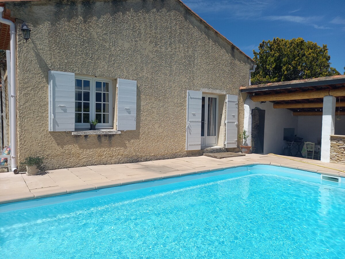 A serene swimming pool is situated in front of a textured stone exterior, with two windows framed by white shutters. The expansive pool area features light-colored tiles, offering a spacious environment for relaxation and enjoyment under clear blue skies.