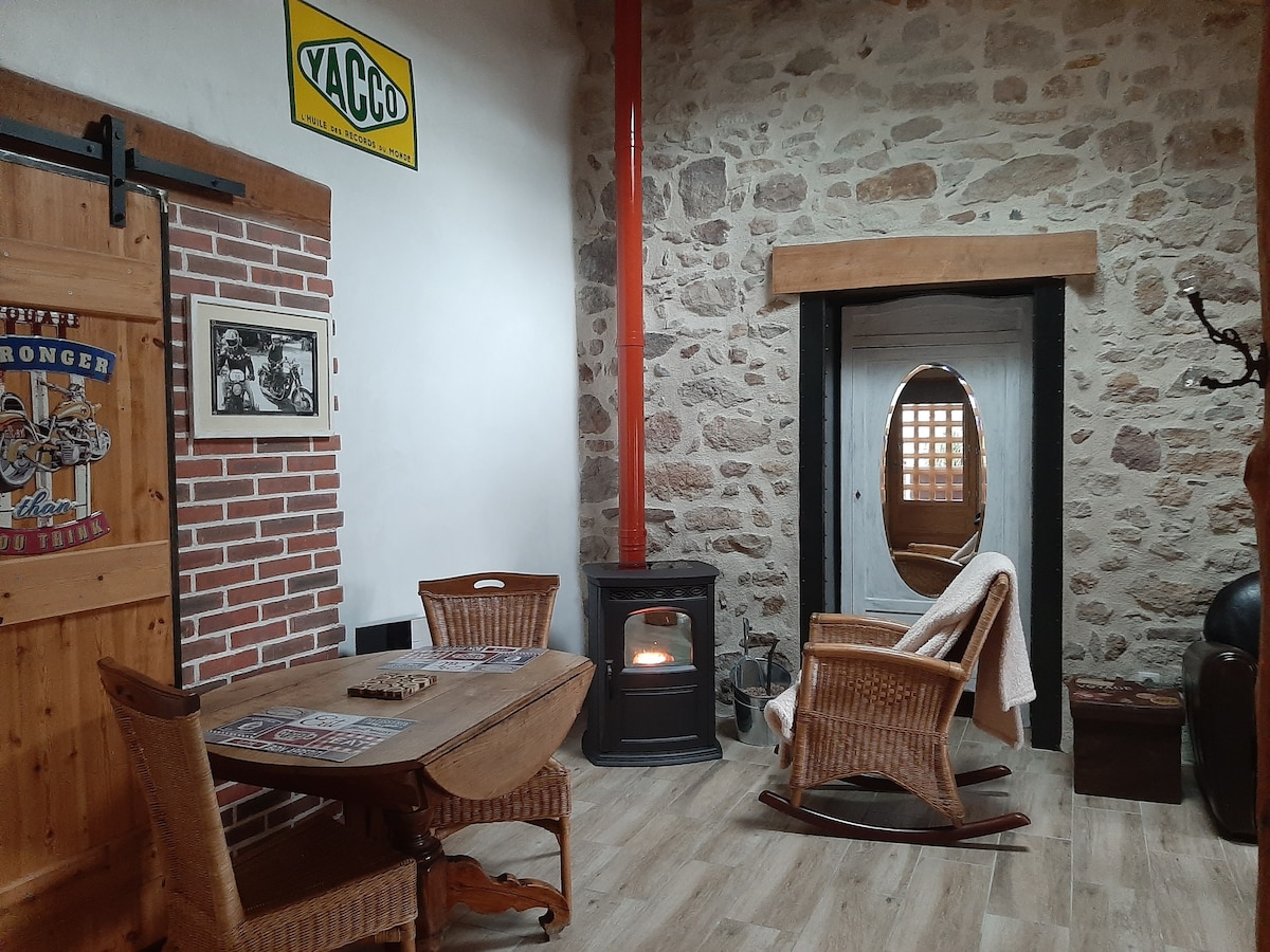 A cozy sitting area features a stone wall and wooden accents. A rustic table is paired with two wicker chairs, while a small wood-burning stove adds warmth. A door with a grid design leads to the outside, and a blanket drapes over one chair.