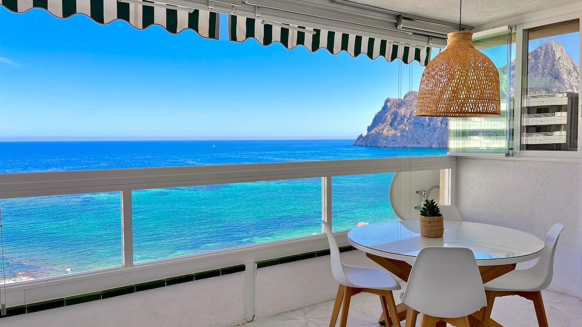 A dining area is presented with a round table surrounded by white chairs, overlooking the ocean. Large windows offer panoramic views of the vibrant blue sea and distant cliffs, while natural light fills the space. A pendant lamp made of woven materials hangs above the table.