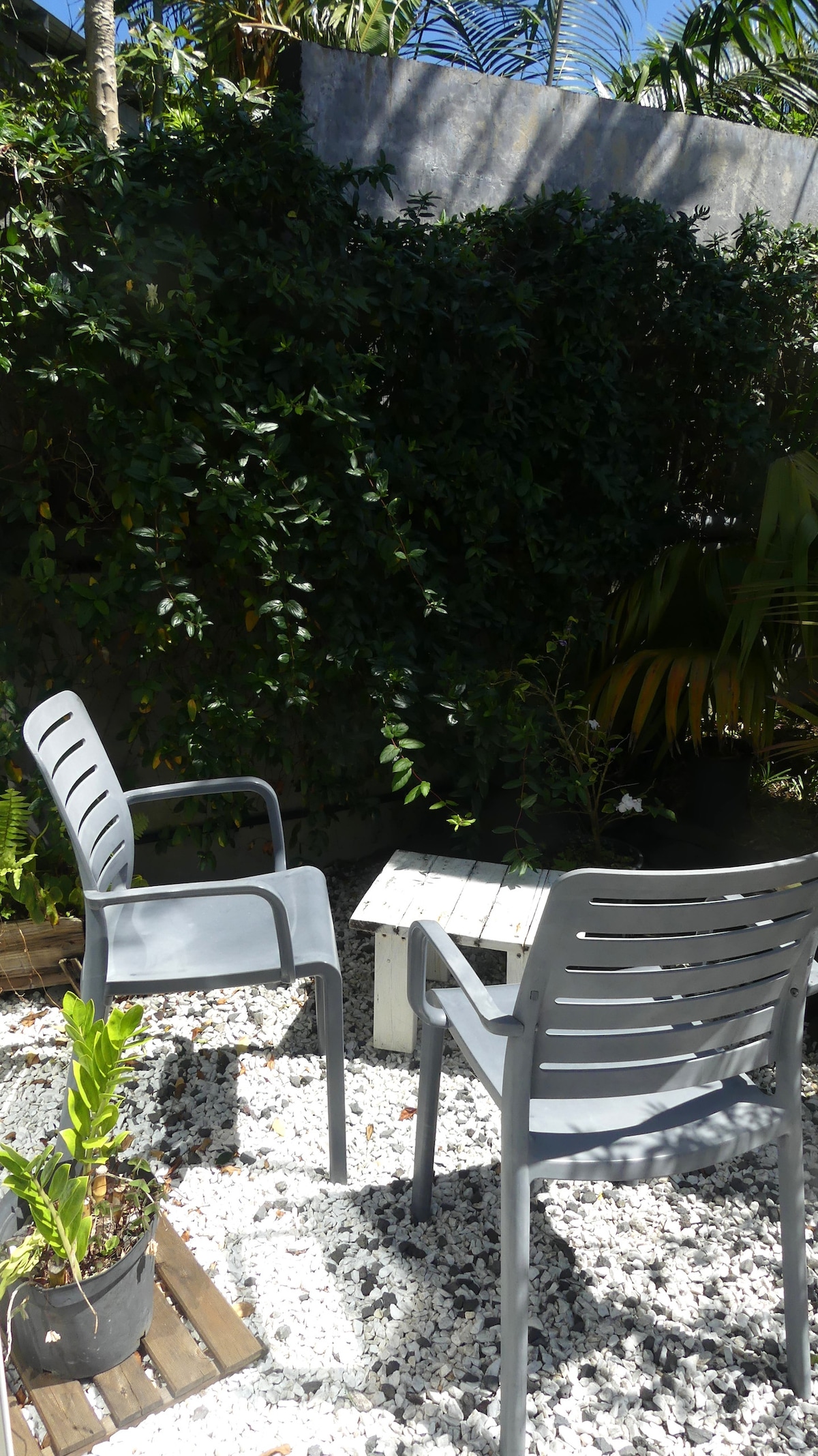 A private outdoor space features two gray chairs positioned around a small table on white gravel. Lush greenery provides a natural backdrop, while potted plants add a touch of color. The area offers a serene setting for relaxation.