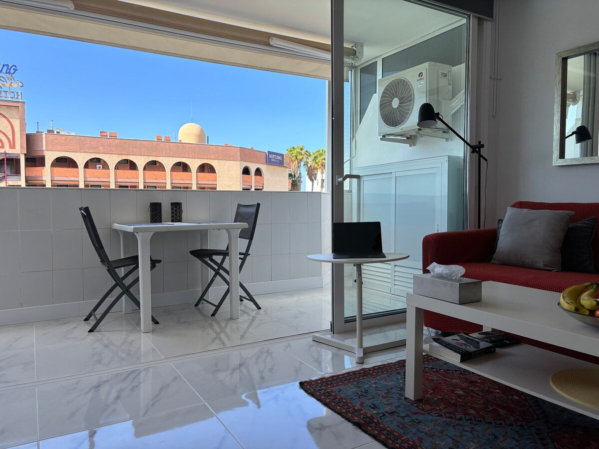 A bright living space is presented, featuring a small dining table with two folding chairs and a modern sofa in red. Natural light floods the area through glass doors, which open to a balcony. A coffee table holds various items, including a bowl of fruit.