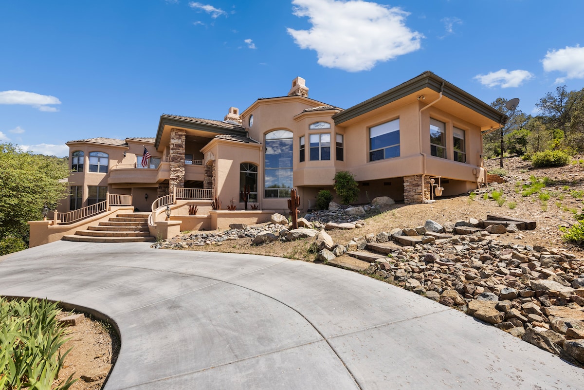 The exterior of the home is showcased, featuring a distinctive architectural design. A curved driveway leads to the entrance, flanked by landscaped areas with boulders and greenery. Large windows offer views of the surrounding landscape, highlighting the serene and inviting atmosphere of the property.