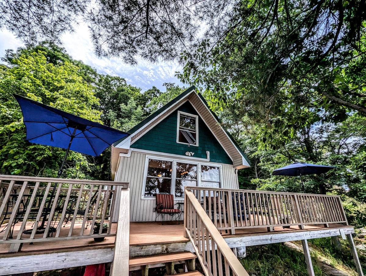 The exterior of the cottage is showcased, featuring a two-level deck with wooden railings. Large blue umbrellas provide shade over seating areas. Lush green trees surround the property, creating a secluded atmosphere, while a triangular gable roof enhances the cabin's charm.