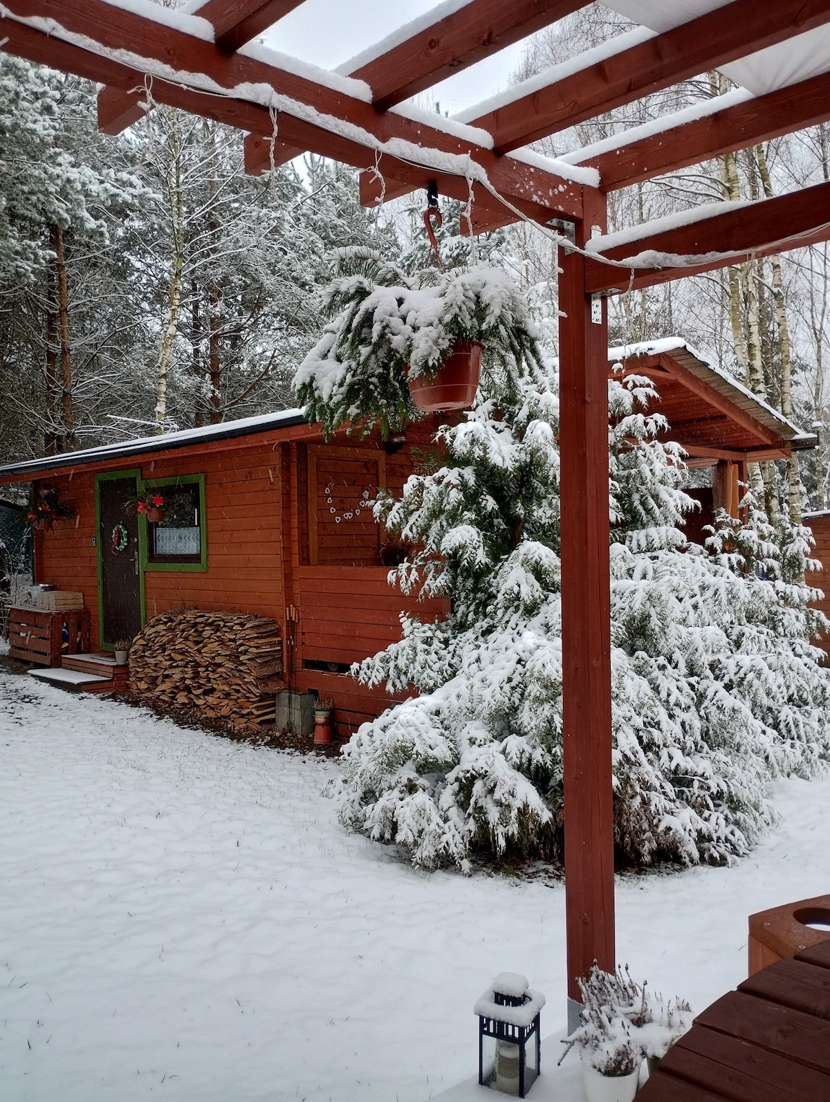 A wooden cabin is nestled among snow-covered trees, with a layer of snow blanketing the ground. Stacked firewood is arranged nearby, and green decorations adorn the cabin's exterior, adding a festive touch. The scene is framed by a wooden pergola overhead.