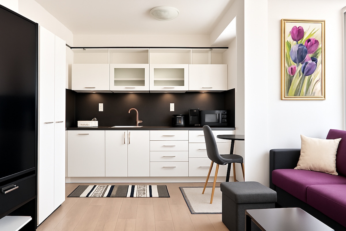 An open-plan kitchen and living area is depicted, featuring modern cabinetry in white and a black countertop. A dining table with a chair is set against the wall, and cozy seating is visible in the living space. A colorful floral artwork adds a touch of vibrancy.