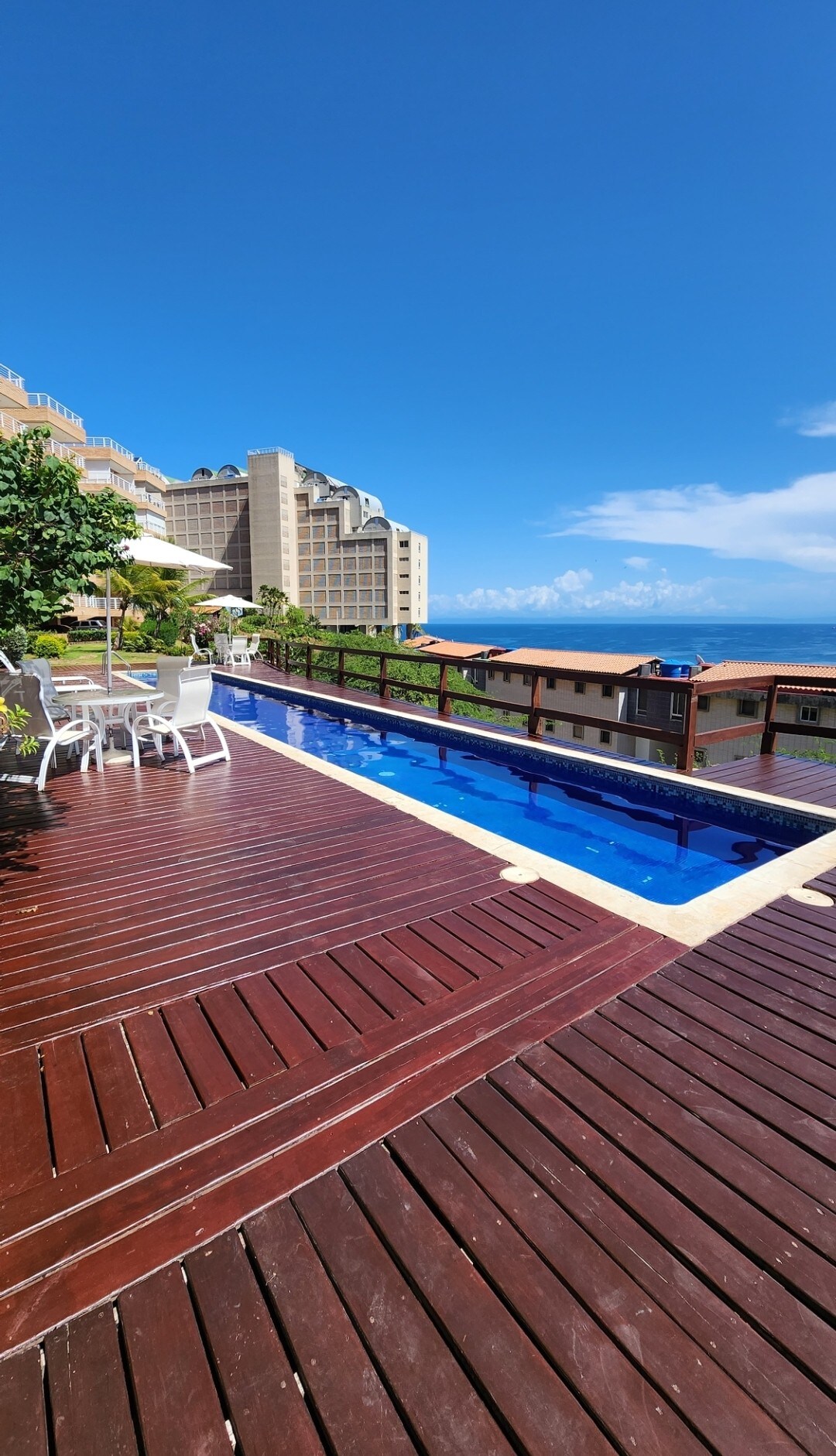 A serene outdoor pool area is presented, featuring a blue swimming pool bordered by wooden decking. White lounge chairs and a table are positioned nearby, set against a backdrop of clear blue skies and distant buildings. Lush greenery adds a touch of nature to the scene.
