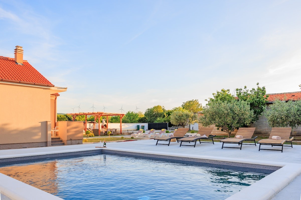 An inviting outdoor space features a rectangular swimming pool surrounded by lounge chairs. The area offers a backdrop of greenery and wind turbines against a clear sky. A covered seating area is visible in the distance, enhancing the tranquil atmosphere.