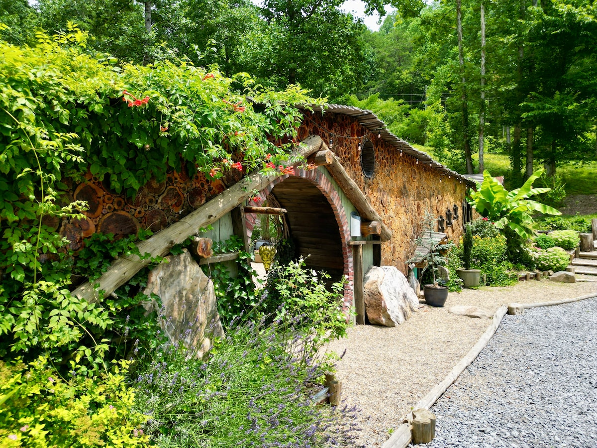 The exterior of the tiny house features unique, textured walls adorned with natural elements. Lush greenery and vines envelop the entrance, which is framed by wooden beams and a stone pathway. The surrounding landscape includes various plants, rocks, and trees, creating a serene environment.