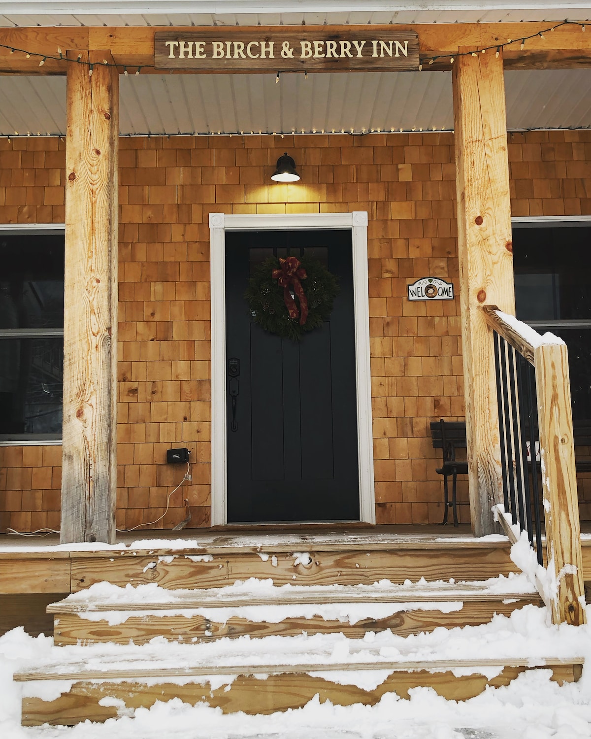A welcoming entrance to the Birch & Berry Inn is highlighted by a wooden porch and warm, rustic wooden shingle siding. Snow covers the steps and ground, while a festive wreath adorns the front door. A sign above the door reads "The Birch & Berry Inn."