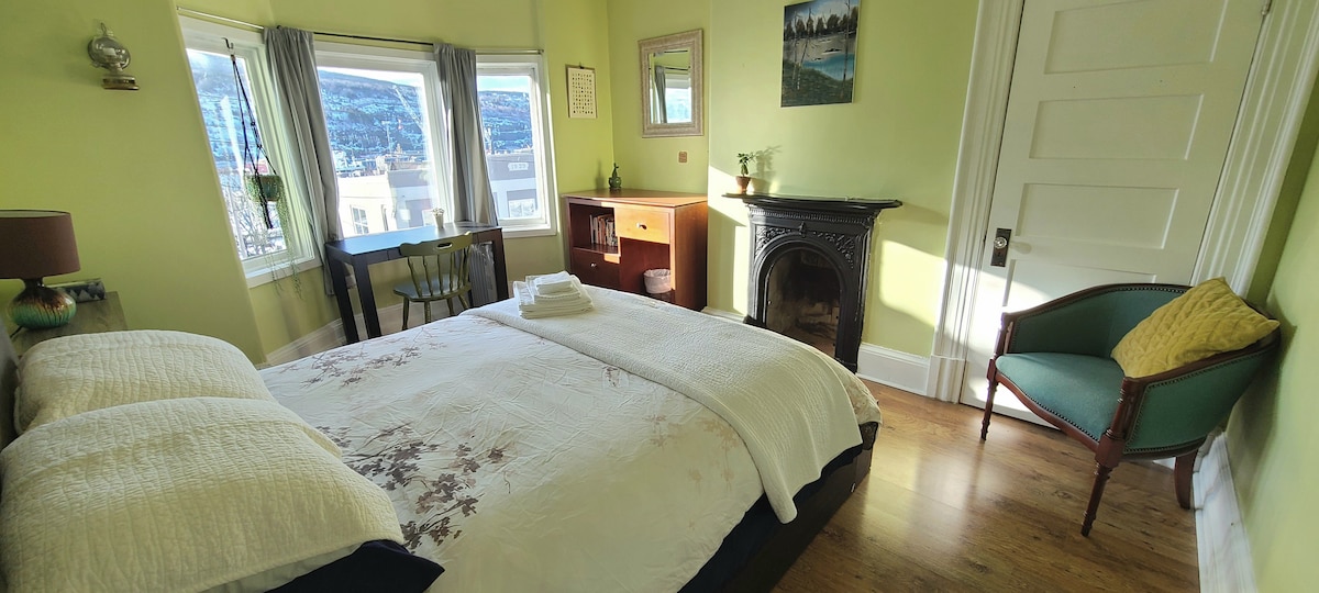 A sunlit bedroom highlights large windows offering a view of the harbour. The spacious bed is adorned with a light blanket, complemented by a small seating area with a green chair. A vintage-style fireplace adds character, while a desk and cabinet provide functional elements.