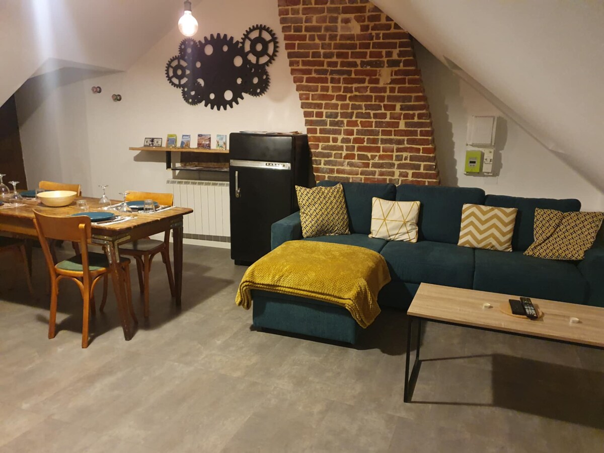 The open living area features exposed brick walls and a cozy seating arrangement with a blue couch adorned with patterned cushions. A wooden dining table is set for four guests, and a black refrigerator is positioned nearby, contributing to the modern atmosphere.