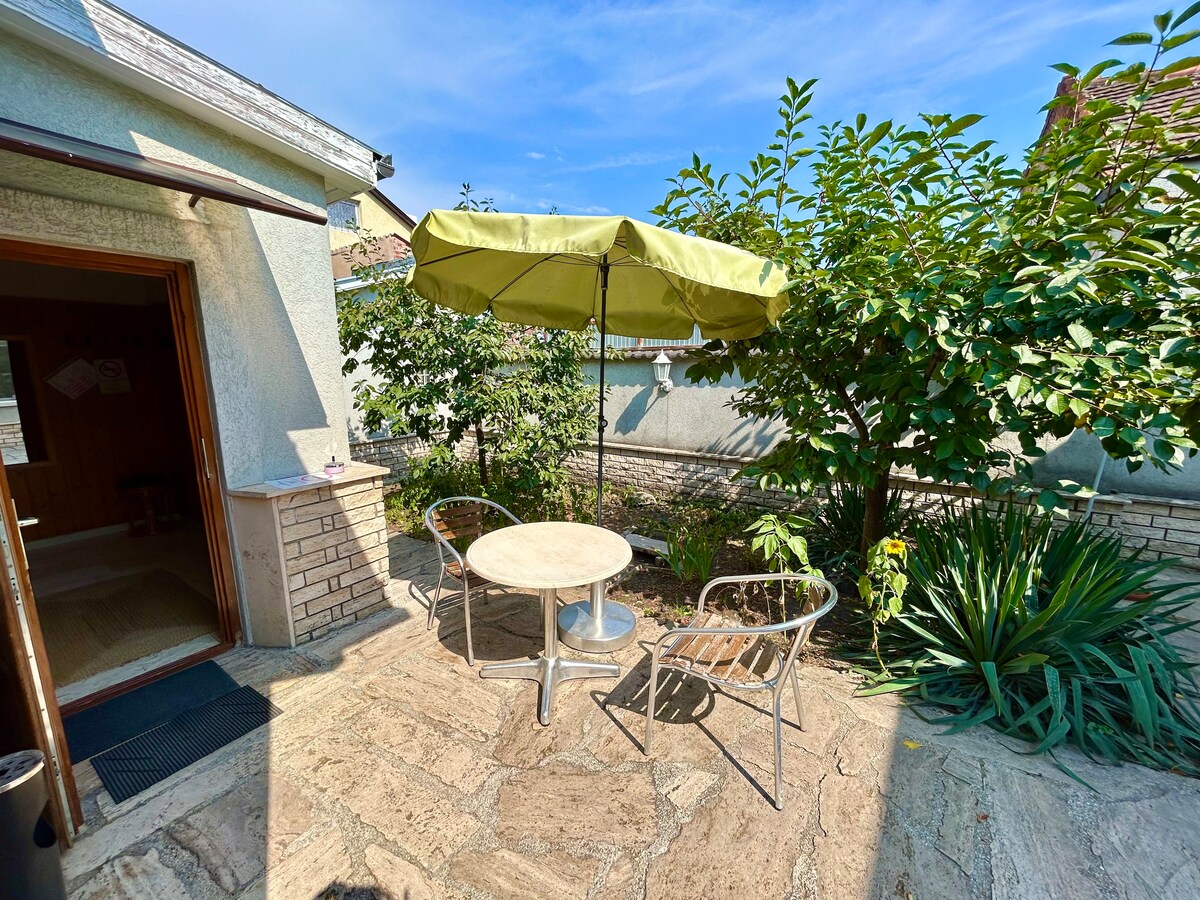A tranquil outdoor space is highlighted, featuring a round table surrounded by two chairs beneath a green umbrella. Lush greenery, including bushes and plants, frames the area, providing a sense of privacy and comfort.