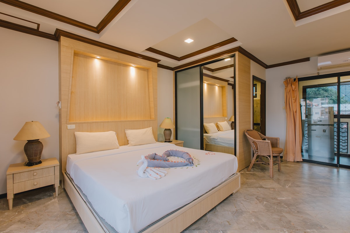 The spacious room features a comfortable bed with neutral bedding, complemented by two bedside lamps. A large mirror reflects the light, enhancing the bright atmosphere. Glass sliding doors lead to an outdoor area, while a seating arrangement adds to the room's functionality.