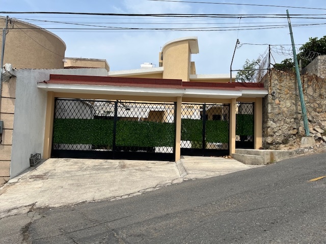 A secure entrance is depicted with a gated driveway bordered by green foliage. The structure features a contemporary design with a sloped roof. The surrounding area includes a sloped street and stone walls, contributing to the residential character.