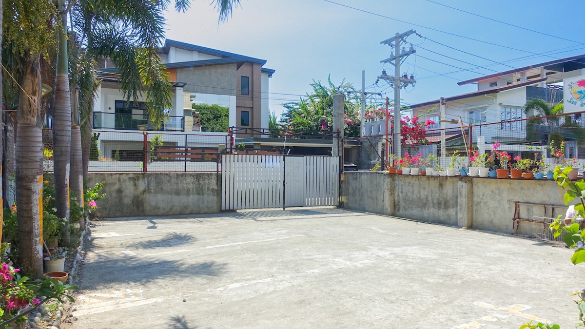 An outdoor parking area is shown, featuring a paved surface with available parking spaces. A gated entrance is visible, surrounded by palm trees and flowering plants in pots. The surrounding buildings provide a glimpse into the neighborhood, ensuring a secure and welcoming environment.