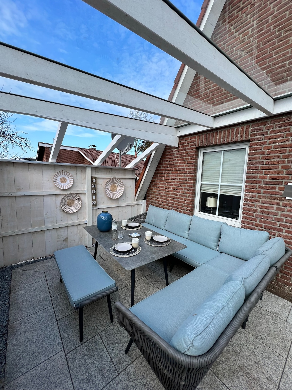 A covered outdoor seating area features a large sectional sofa with light blue cushions, complemented by a matching bench and a gray dining table. Decorative wall elements are visible along the wooden fence, and the area is bathed in natural light from the clear sky above.