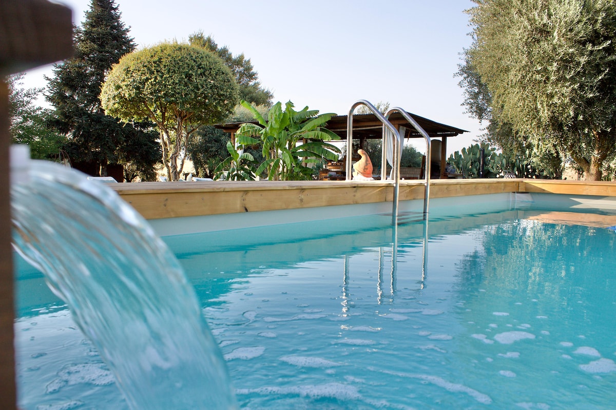A modern swimming pool is set within a wooden deck, featuring a gentle waterfall feature. Lush greenery surrounds the pool, with tall trees and shrubs visible. A shaded gazebo can be seen in the background, providing a relaxed outdoor setting.