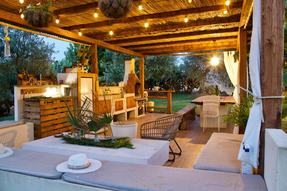 A spacious gazebo features comfortable seating areas and an outdoor kitchen, providing a shaded space for relaxation. Soft lighting creates a cozy ambiance. The surrounding greenery enhances the tranquil environment, complemented by a nearby hammock and barbecue facilities.
