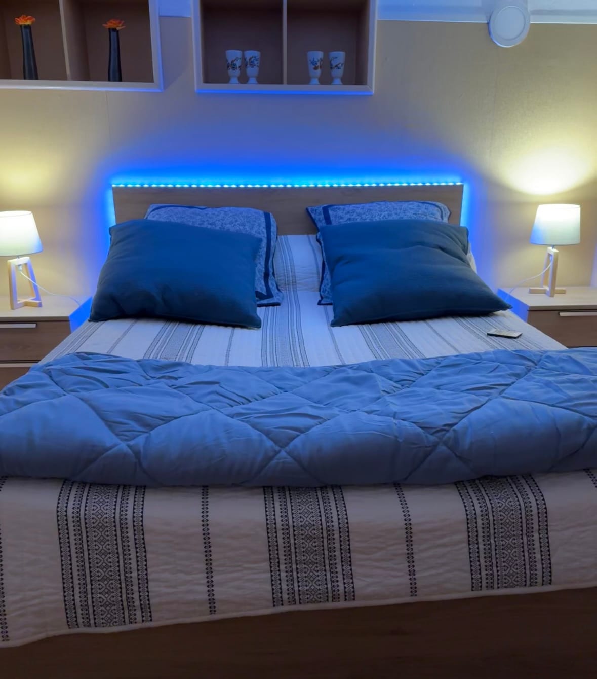 A spacious bed is presented with layered blue and white bedding, accented by two large pillows. Soft lighting is provided by two bedside lamps, creating a gentle ambiance. A decorative shelf above features minimalist decor items, adding a touch of character to the room.