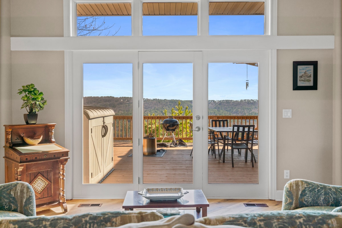 A spacious living area showcases large glass doors that open to a deck with panoramic mountain views. A grill is visible on the deck, and a dining set offers outdoor seating. The interior features a comfortable sofa and a decorative table, creating a welcoming environment.