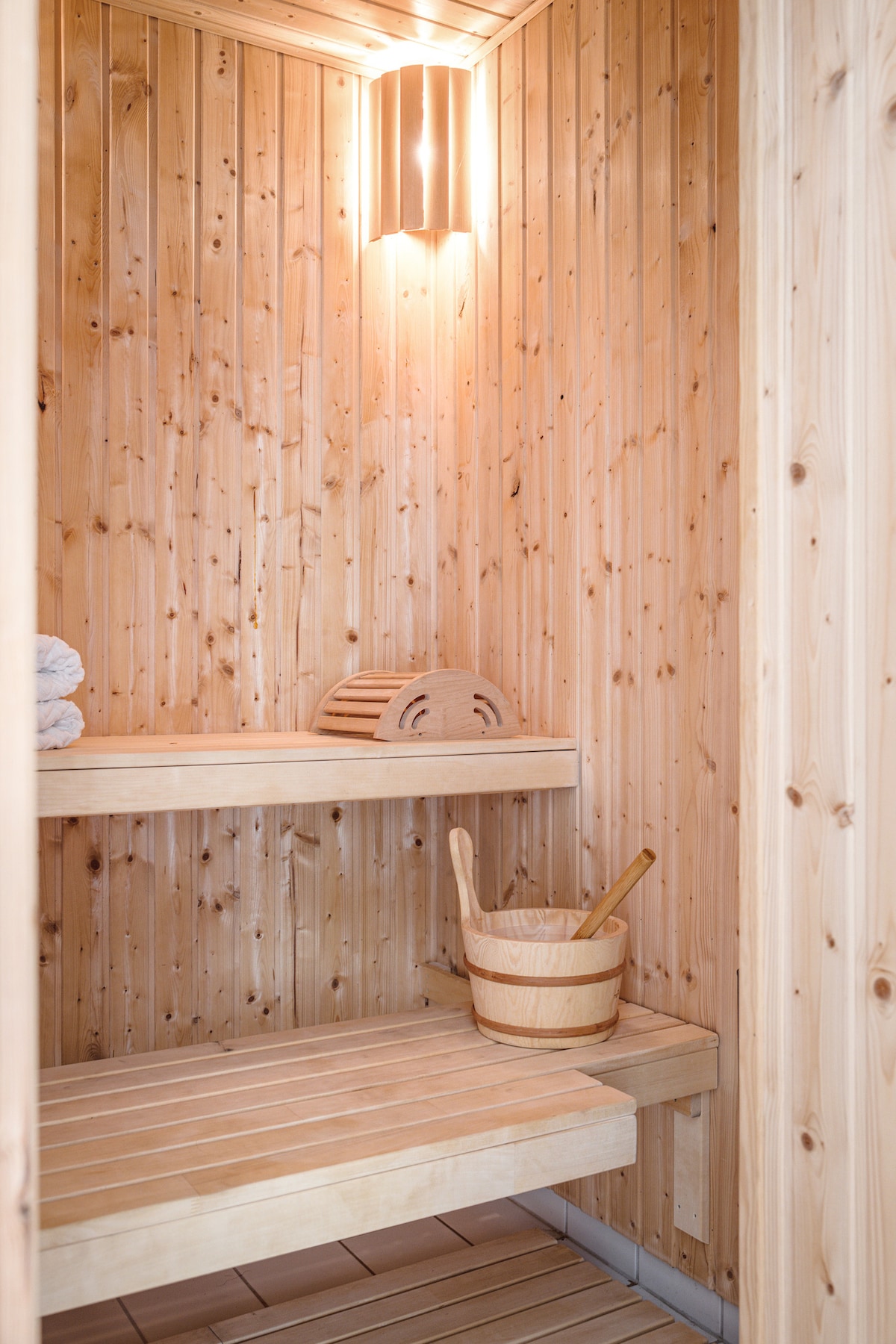 A wooden sauna interior is displayed, featuring warm-toned planks and soft lighting. Benches are positioned on two levels, while a wooden bucket and ladle sit invitingly on a lower shelf. A stack of towels adds a touch of convenience for guests.