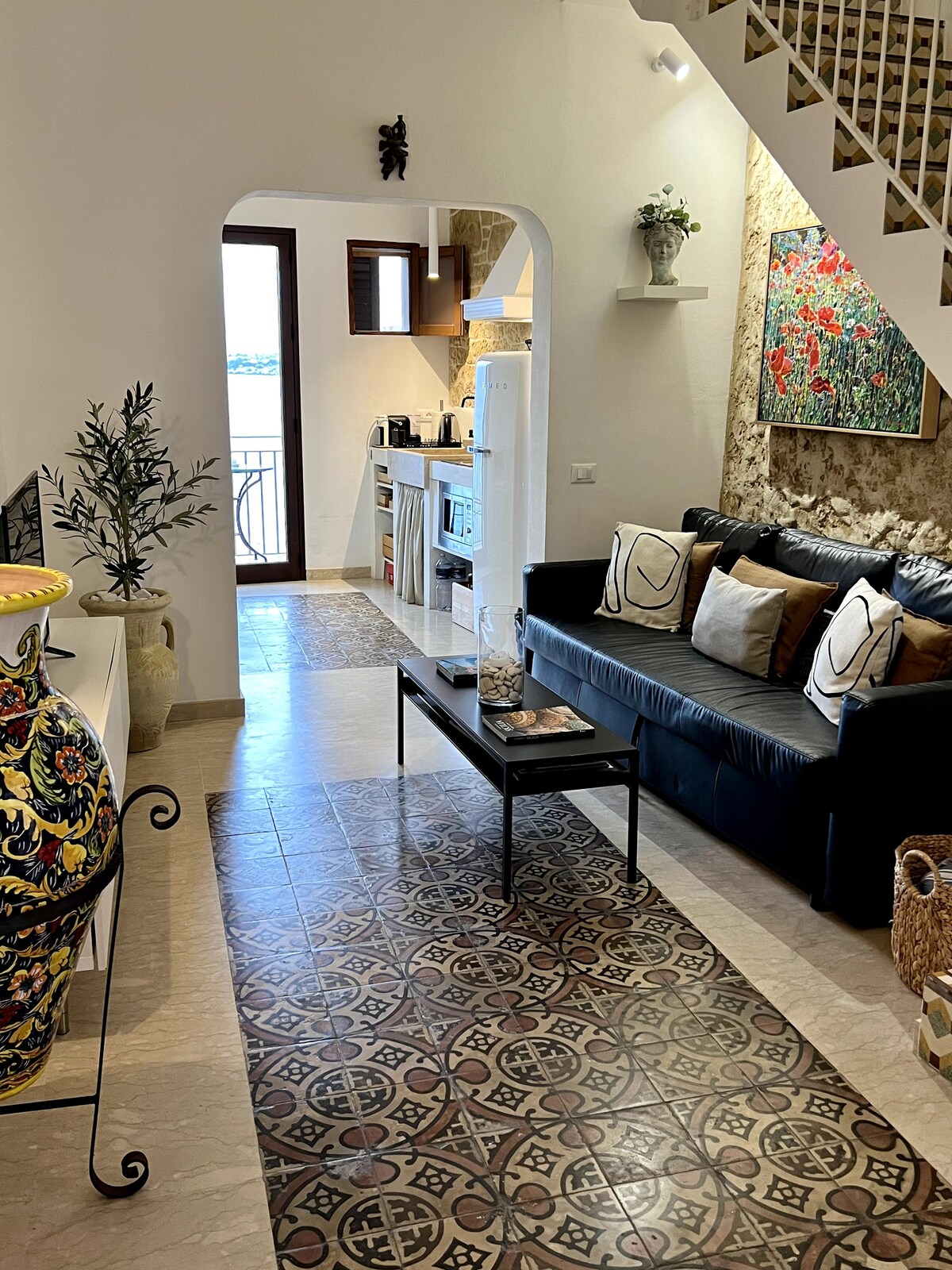 A spacious living area features a black sofa adorned with decorative pillows, complemented by a sleek coffee table. Bright light filters through a window, illuminating the colorful art piece on the wall. A charming tiled floor leads towards the kitchen area, enhancing the welcoming ambiance.