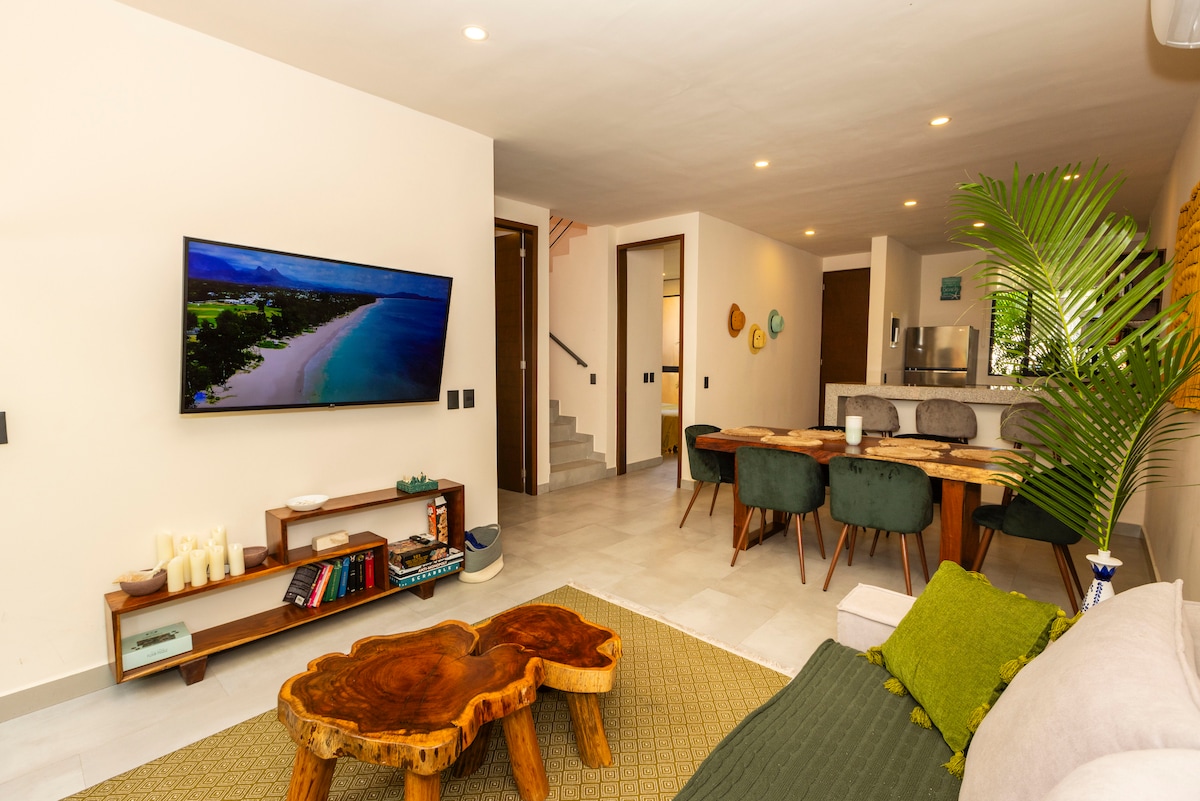 An open-concept living area features a comfortable sofa and a wooden coffee table, with a large HD Smart TV mounted on the wall. A dining table with upholstered chairs is visible, as well as access to the kitchen area, creating a spacious and inviting environment.