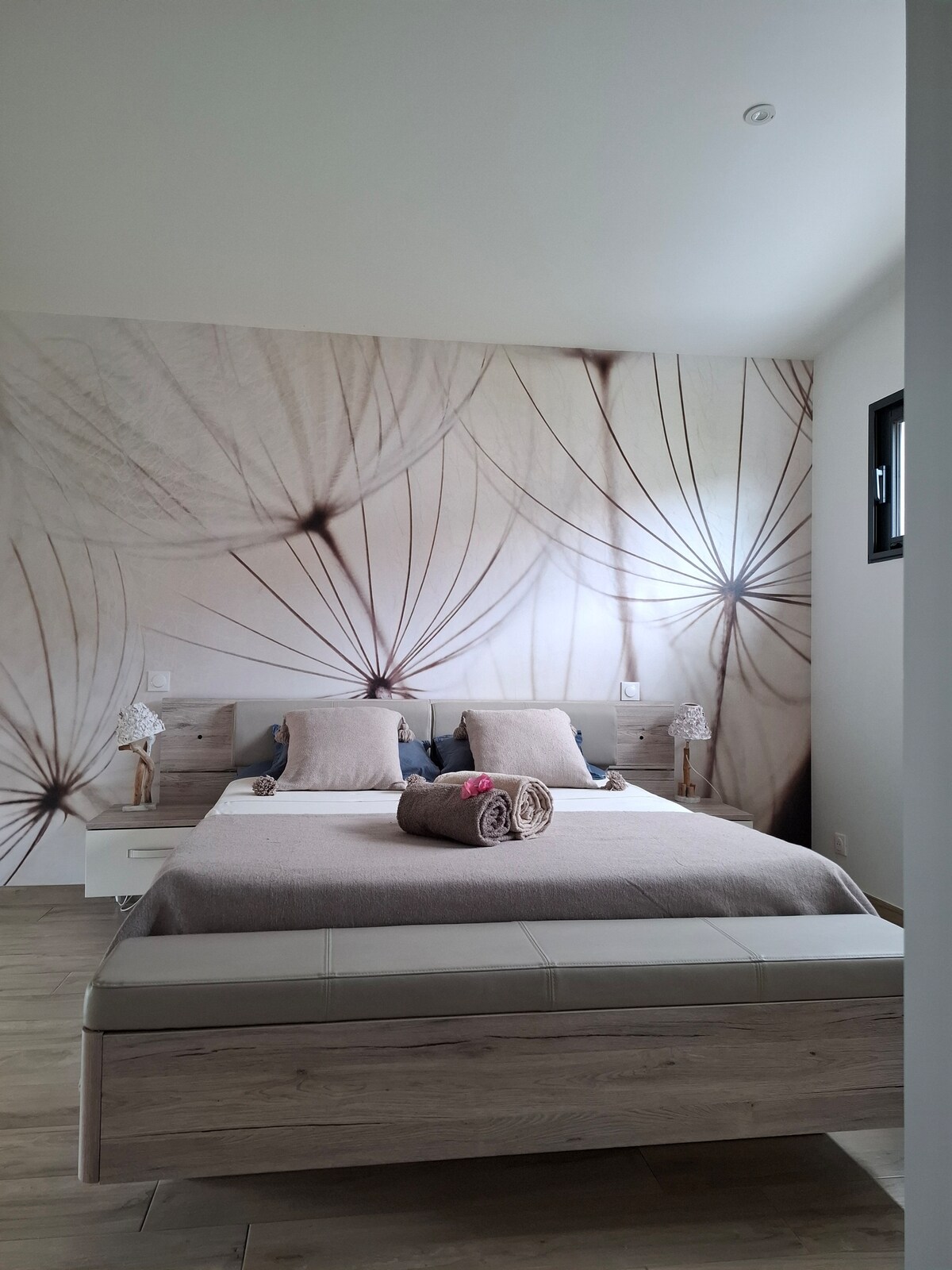 The bedroom features a spacious bed with a neutral-colored blanket and decorative pillows. Soft light illuminates the room, highlighting a wall adorned with a delicate dandelion design. Nightstands are placed on either side of the bed, each with a lamp.