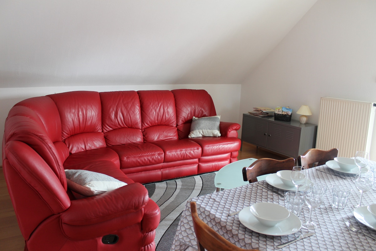 A spacious seating area features a large red leather sofa accompanied by a small decorative pillow. A dining table is set with dishware and glassware, while a sideboard provides additional storage space. Natural light is filtered gently through the room's designed elements.