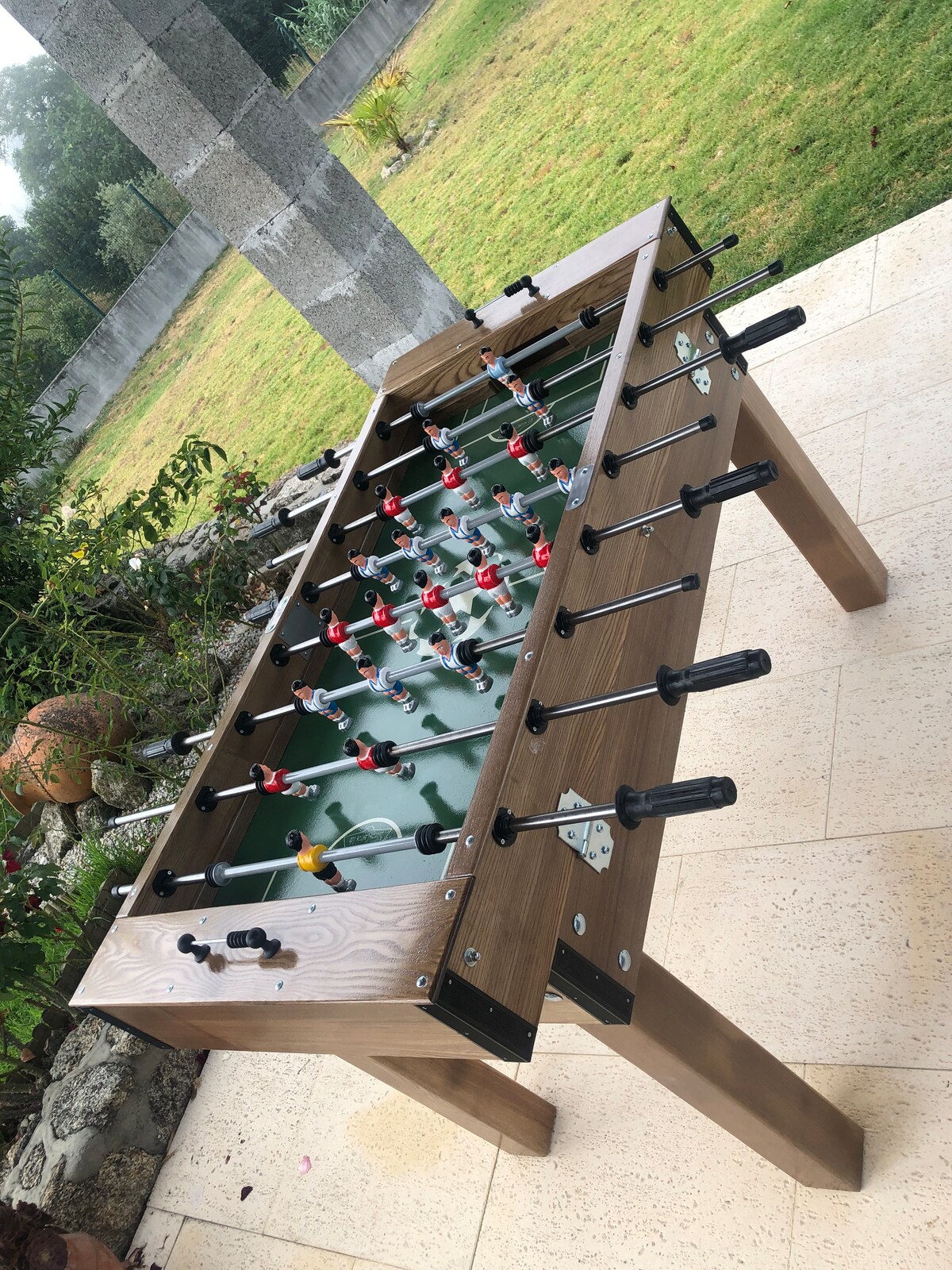 A foosball table is positioned outdoors on a patio, surrounded by greenery. The table features a wooden finish with black handles and colorful player figures, inviting engagement and leisure. The grassy area in the background provides a natural setting for relaxation and fun.