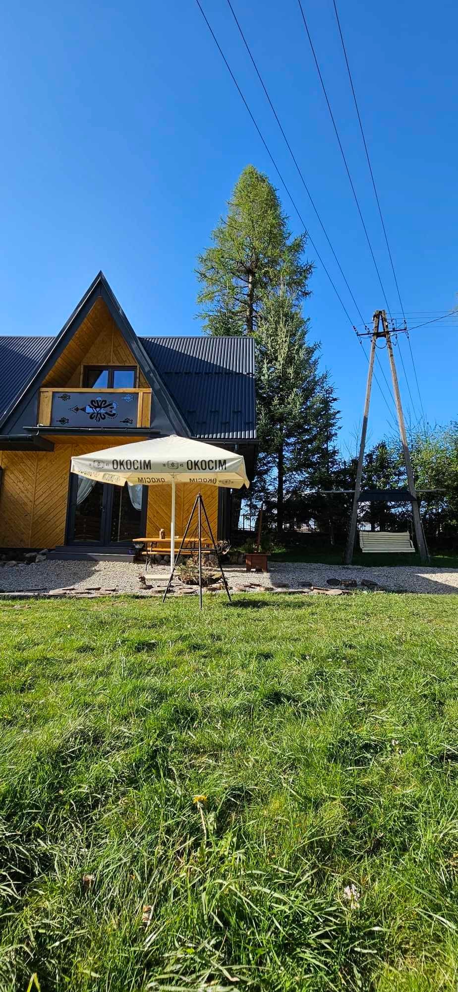 The exterior of a wooden cabin is shown, featuring a sloped roof and balcony. A swing hangs beneath a canopy, framed by green grass. Towering trees are visible in the background, while utility poles stand along the edge of the property.