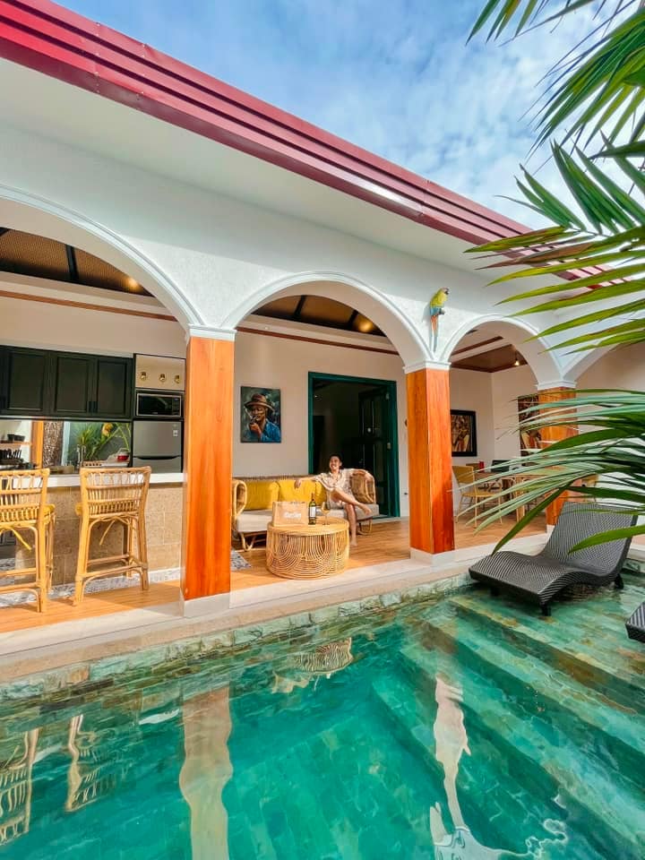 A tropical bungalow opens to a private pool, featuring arched doorways and a cozy seating area. Sun loungers are placed alongside the crystal-clear water, with lush greenery surrounding the space. Inside, a well-equipped kitchen is visible, enhancing the inviting atmosphere.