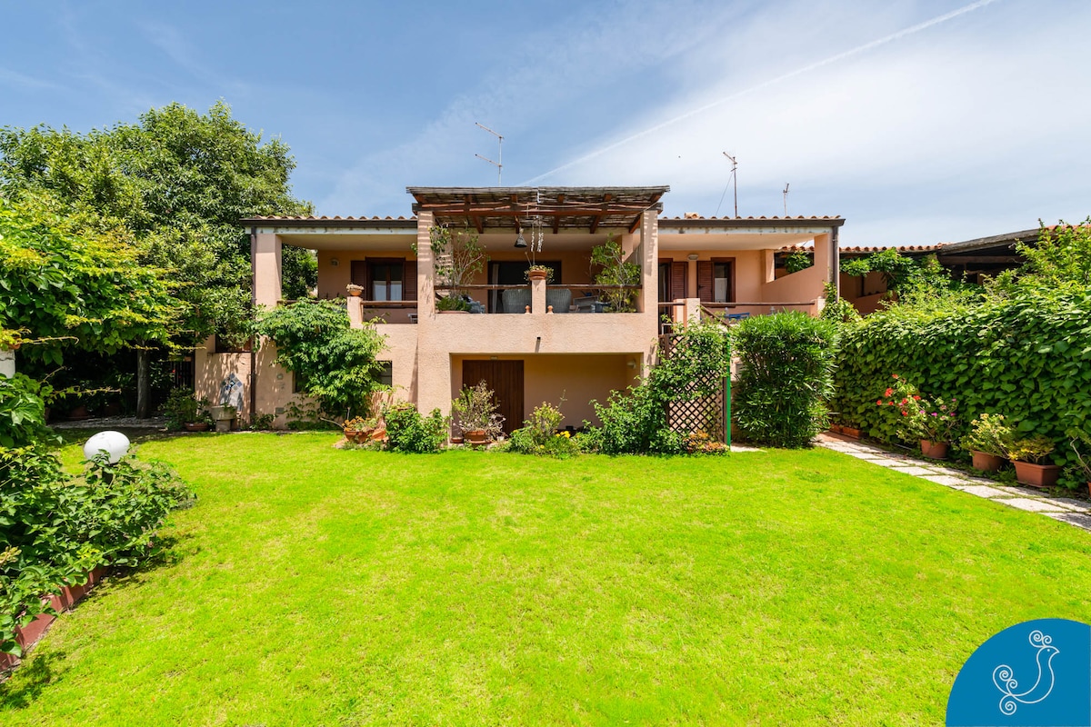 The villa is surrounded by a lush green garden, featuring a spacious lawn and numerous flower beds. Two patios are visible on the upper level, framed by elegant pillars. The property is enclosed by vibrant greenery, providing privacy and a serene outdoor environment.