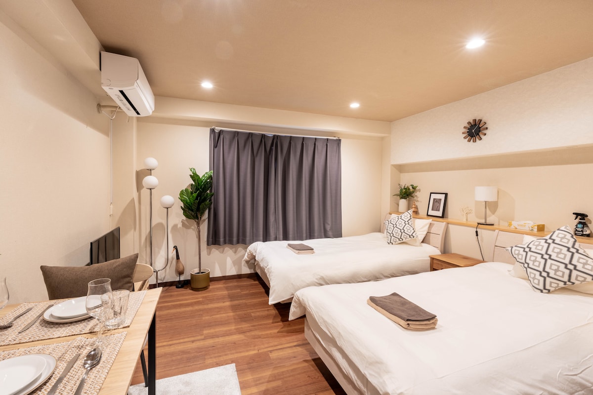 The room features two spacious beds positioned side by side, accented by modern decor. A dining area with a table and place settings is visible. A potted plant and standing lamp add touch points, while a large window with dark curtains allows for natural light.