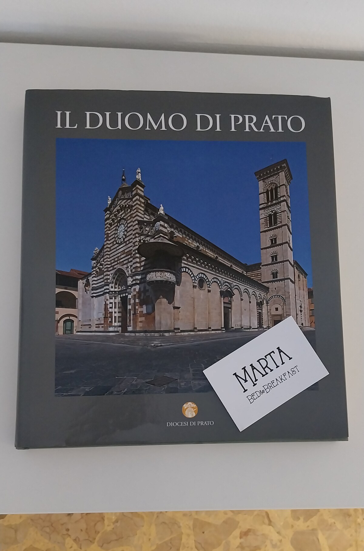 A hardcover book titled 'Il Duomo di Prato' is displayed on a flat surface. The book features a striking image of the Duomo, showcasing the building's architectural details against a clear blue sky. A label with the name 'Marta' is attached to the book's cover.