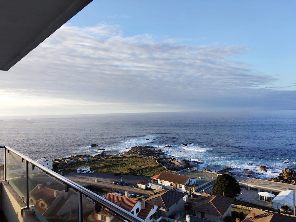 A serene coastal view is captured from a balcony, showcasing the expansive blue ocean and rocky shoreline. Rolling waves meet the land, while a clear sky with scattered clouds enhances the tranquil setting. The surrounding area features charming homes along the coast.