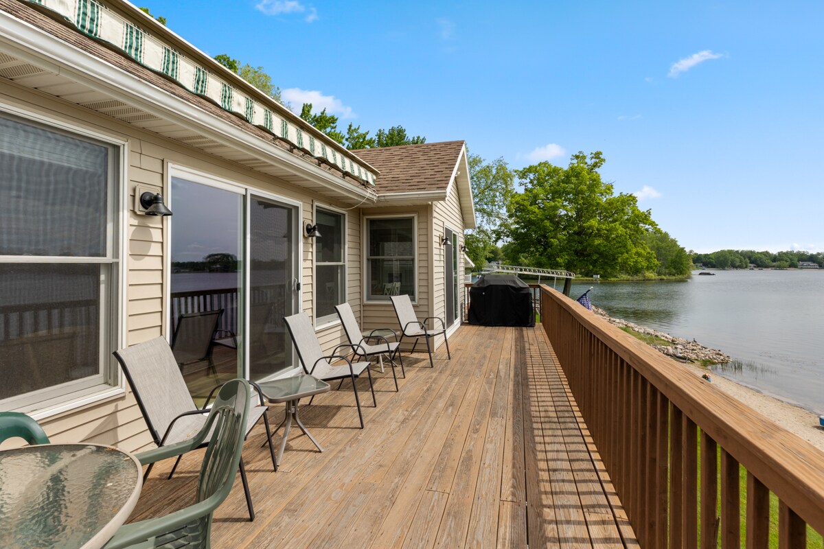 A spacious deck offers multiple seating options, including four lightweight chairs and a glass table. The views of the lake and surrounding greenery are visible, with the water's edge just a short distance from the deck.
